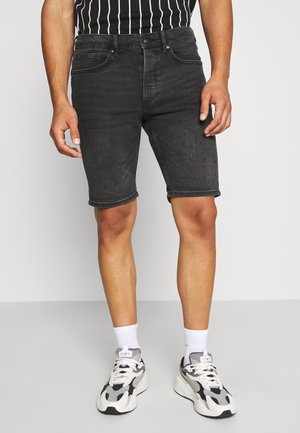 Mann trägt schwarze Jeansshorts, schwarz-weiß gestreiftes Hemd, weiße Socken und weiße und schwarze Turnschuhe, steht auf grauem Boden.