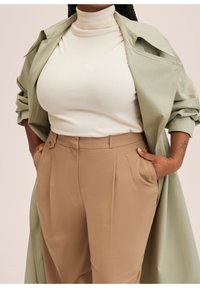 Un pull col roulé crème sous un trench-coat vert clair, associé à un pantalon beige taille haute avec poches latérales et plis.