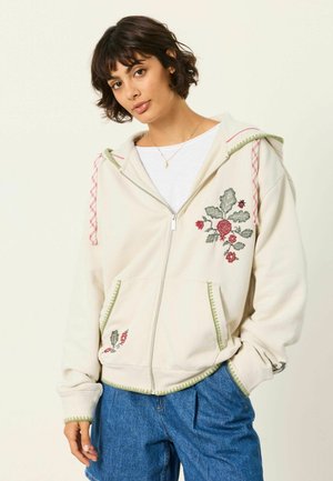 Kvinde med kort mørkt hår iført en cremefarvet hættetrøje med lynlås og blomsterbroderi samt grøn kant, kombineret med blå denimshorts.