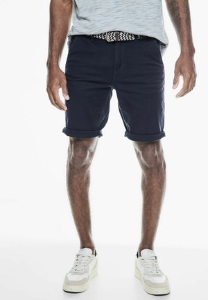 Navy blue shorts with rolled hems, made of cotton. Worn with a light gray shirt and a patterned belt, paired with white sneakers.