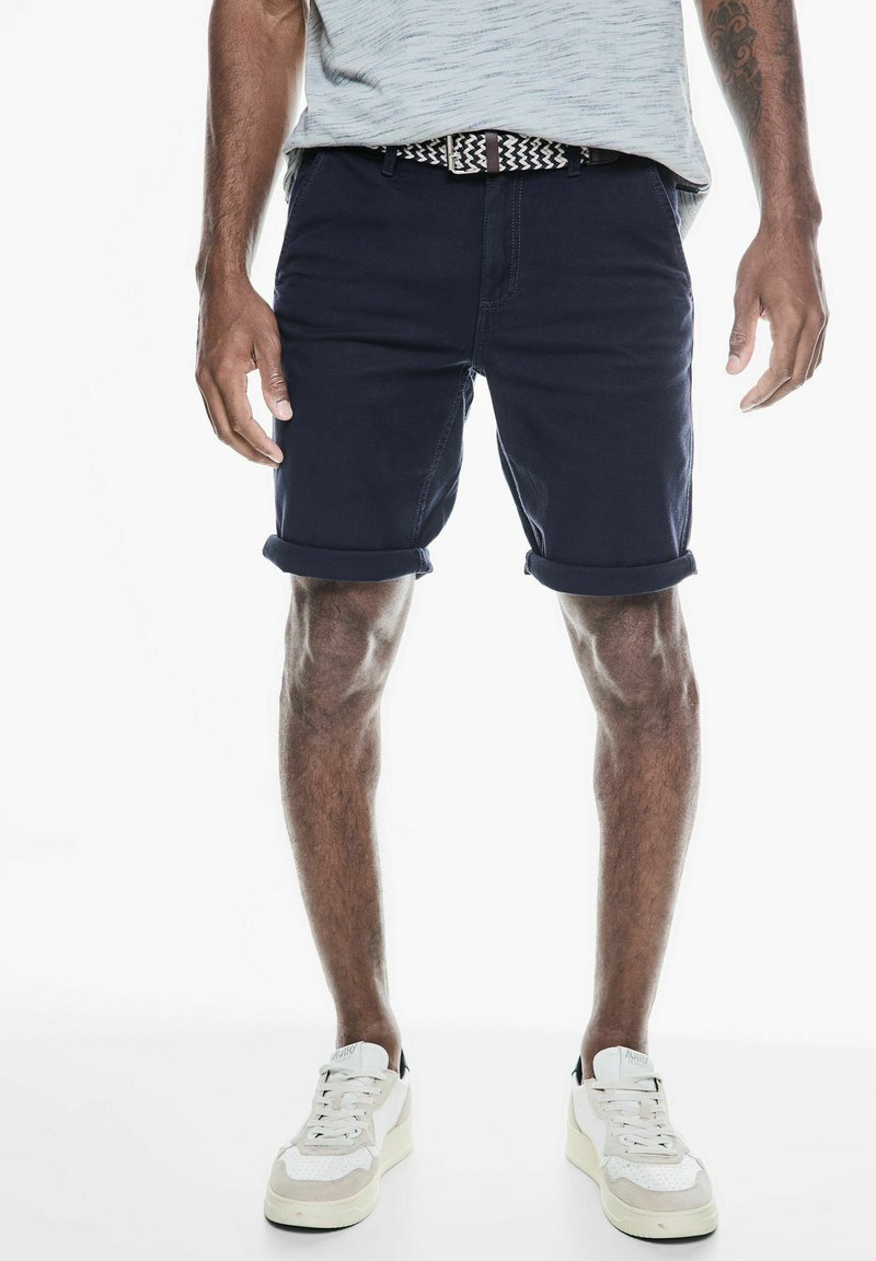 Marineblauwe shorts met opgerolde zomen, gemaakt van katoen. Gekocht met een lichtgrijs shirt en een patroonriem, gecombineerd met witte sneakers.