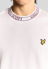 Weißes T-Shirt mit kurzen Ärmeln und Rundhalsausschnitt, das mit einem kastanienbraunen Band versehen ist. Enthält ein kleines, gelbes, gesticktes Vogel-Logo auf der Brust.