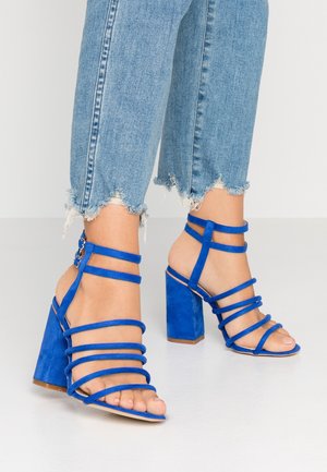 Voeten met blauwe sandalen met bandjes en hoge hak, gecombineerd met blauwe denimjeans met rafelige zoom op een lichte vloer.