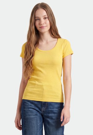 Jonge vrouw met lang lichtbruin haar, gekleed in een geel shirt met korte mouwen en blauwe spijkerbroek, staand tegen een witte achtergrond.
