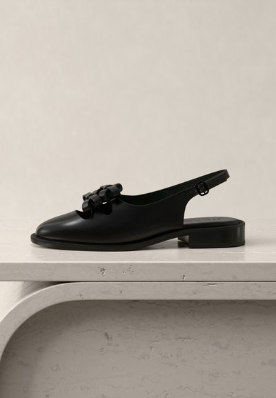 Black leather slingback flat shoe with low heel and decorative bows on the front, displayed on a light textured surface.
