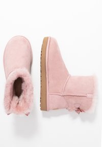 Bottines en daim rose clair avec doublure en fausse fourrure, bout arrondi et semelle en caoutchouc marron. Comprend un nœud décoratif au talon.