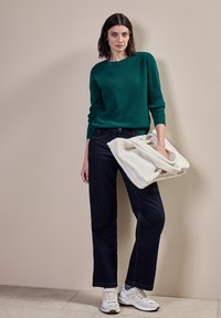 Pull en maille côtelée verte, jeans droit bleu marine, sac en peluche blanc et baskets blanches avec des accents gris. Textures lisses et couleurs contrastées.