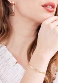 Goldenes Armband mit einem rechteckigen Namensschild und Kettenmuster. Wird am Handgelenk getragen, kombiniert mit hängenden Ohrringen. Weiße strukturierte Bluse.