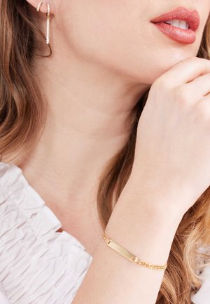Goldenes Armband mit einem rechteckigen Namensschild und Kettenmuster. Wird am Handgelenk getragen, kombiniert mit hängenden Ohrringen. Weiße strukturierte Bluse.