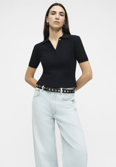 Γυναίκα με μακριά σκούρα μαλλιά που φοράει μαύρο κοντομάνικο πουκάμισο με γιακά, ανοιχτό μπλε τζιν με φαρδιά πόδια και μαύρη ζώνη με τρουκς, στέκεται με τα χέρια πίσω από την πλάτη.