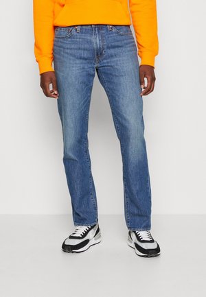 Homme portant un jean bleu droit, des baskets noir et blanc, et un haut orange à manches longues sur un fond uni.