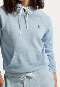 Nainen, joka pitää yllään vaaleansinistä hupparia ja siihen sopivia verkkareita, joissa on valkoiset nyörit ja pieni tummansinisestä langasta brodeerattu logo rinnassa ja vyötäröllä.