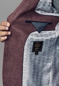Veste bordeaux à l'intérieur avec une doublure géométrique argentée. Comprend une étiquette indiquant "NEXT, Coupe Ajustée" et un rabat de poche bleu marine.