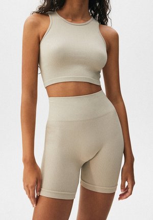 Nainen, joka pukeutunut beigeen, ribbineulottuun, hihattomaan crop-toppiin ja korkeavyötäröisiin ribbineulottuihin pyöräilyshortseihin, seisoo yksiväristä taustaa vasten.