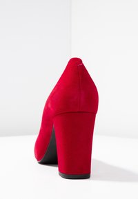 Czerwone zamszowe buty na wysokim obcasie z okrągłym noskiem i szerokim czarnym obcasem. Gładka tekstura i bezszwowy design podkreślają ich odważny wygląd.