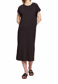 Femme portant une robe midi noire unie à manches courtes et fentes sur les côtés, associée à des sandales plates noires à lanières, se tenant les mains derrière le dos.