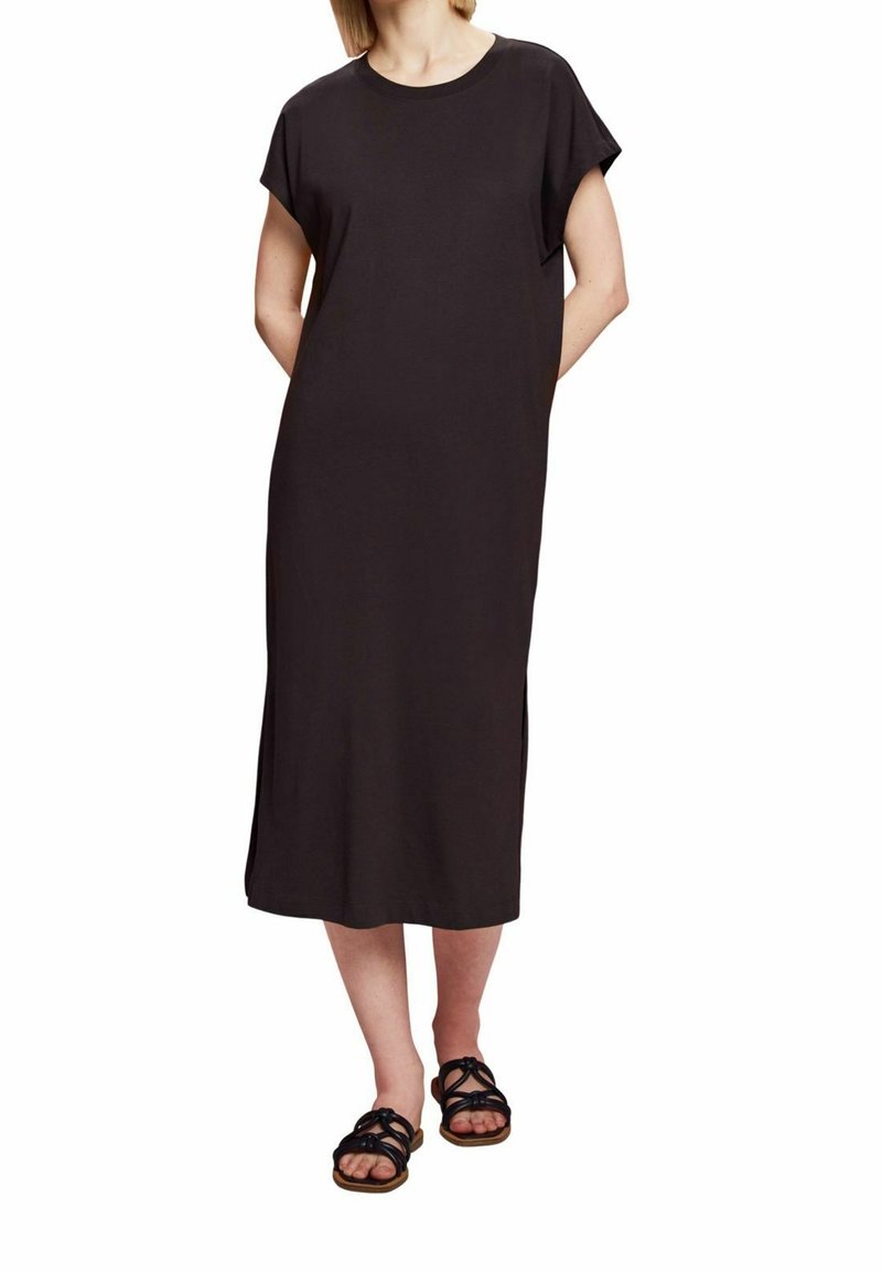 Femme portant une robe midi noire unie à manches courtes et fentes sur les côtés, associée à des sandales plates noires à lanières, se tenant les mains derrière le dos.