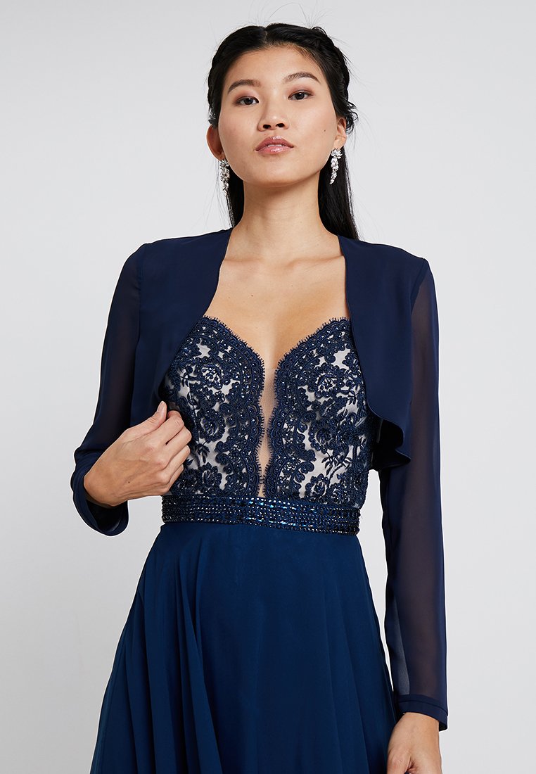 Abito da sera blu navy con maniche lunghe trasparenti, scollatura a V profonda decorata con pizzo e perline, e una gonna fluente. Design elegante e dettagli intricati.
