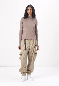 Langärmliges taupefarbenes Oberteil mit Stehkragen; beige Cargo-Hosen mit Reißverschlüssen und elastischen Bündchen; weiße Sneakers. Model steht vor einem schlichten Hintergrund.