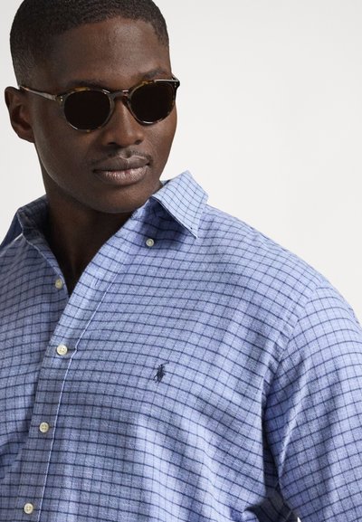 Koszula na guziki w jasnoniebieskim kolorze z wzorem w kratę, wykonana z materiału o miękkiej fakturze. Posiada małe logo na piersi i kołnierz typu rozłożysty. Brązowe okulary przeciwsłoneczne.