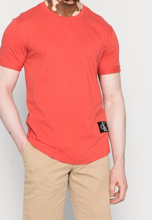 Mann trägt ein schlichtes rotes T-Shirt mit kurzen Ärmeln, mit einem kleinen schwarzen CK-Logo-Patch nahe dem Saum, und beige Shorts, die Hand in der Tasche.