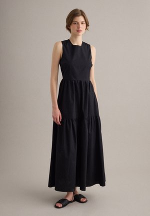 Joven mujer con un vestido maxi negro sin mangas con volantes y sandalias planas negras tejidas, de pie contra un fondo beige liso.