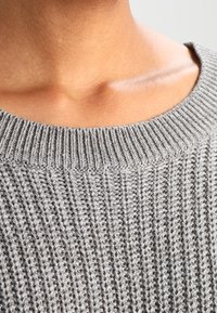 Grå stickad tröja med ribbat mönster och bred rund halsringning, som visar detaljerad sömnad och subtila variationer i garnets tjocklek.