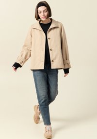 Beżowa wodoodporna kurtka z szerokim kołnierzem, z przodu zapinana na guziki i z podwiniętymi rękawami, w komplecie z niebieskimi dżinsami i jasnymi butami.