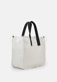 Sac fourre-tout rectangulaire blanc avec des poignées en cuir noir, corps en tissu structuré et un design épuré, posé sur une surface blanche.