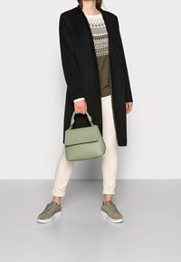 Manteau long noir, pull à motifs olive, pantalon crème, sac à main vert avec anse torsadée, et baskets grises avec semelles blanches.