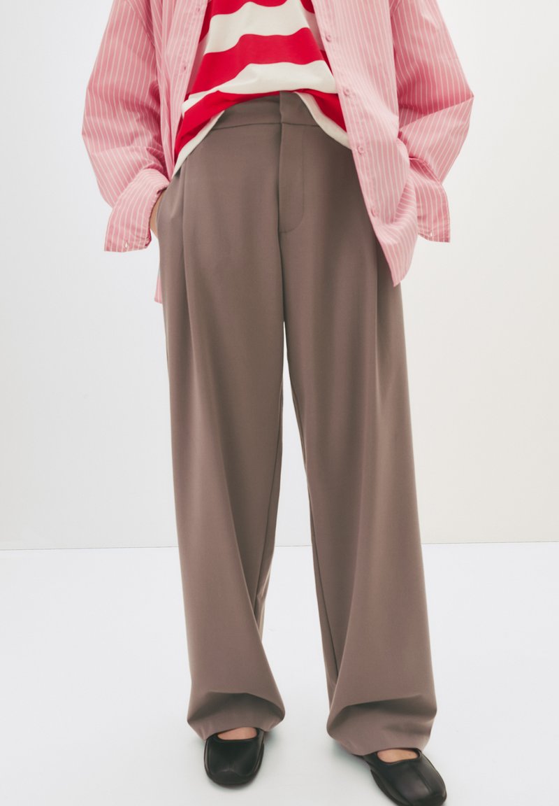 Vide bukser i en blød brun stof, der har en glat tekstur og diskrete folder, matchet med en pink stribet skjorte med knapper.
