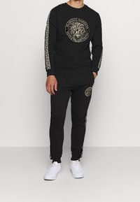 Completo athleisure nero con grafica di un leone sulla felpa, dettagli dorati e pantaloni jogger abbinati con strisce laterali e logo.
