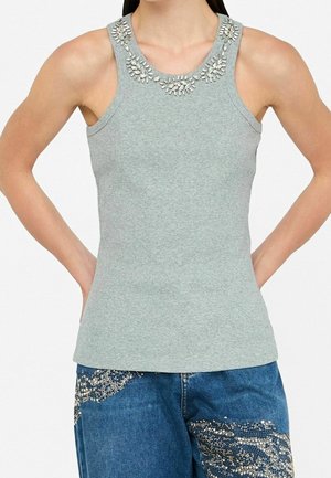 Femme portant un haut gris côtelé sans manches avec un col orné de bijoux et un jean bleu décoré de motifs en strass argentés.
