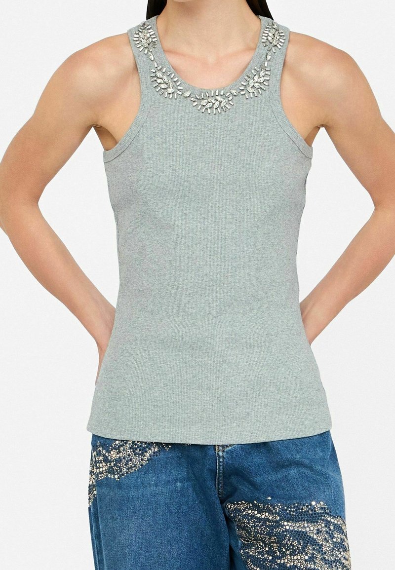 Femme portant un haut gris côtelé sans manches avec un col orné de bijoux et un jean bleu décoré de motifs en strass argentés.