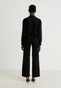 Blouse noire en dentelle à manches longues avec des poignets à boutons, associée à un pantalon large taille haute noir. Des sandales à talons noires viennent parfaire le look.