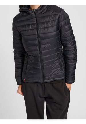 Personne portant une veste noire matelassée zippée avec capuche et un pantalon noir, debout les mains dans les poches sur un fond uni.