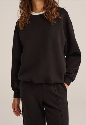 Femme portant un sweat-shirt noir à manches longues et un pantalon noir assorti, une main dans la poche, sur un fond uni.