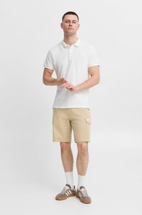 Hvit polo skjorte med kontrastkrage, beige cargo shorts, grå sneakers med hvite striper, og hvite sokker. Stående posisjon.
