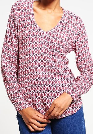 Kvinde iført en langærmet V-hals skjorte med pink og rødt blomsterprint, kombineret med blå jeans, hænderne foldet ved taljen.