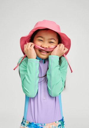 Young girl in colorful long-sleeve swimwear and pink sun hat holding hat strings over her smiling face against a plain background.