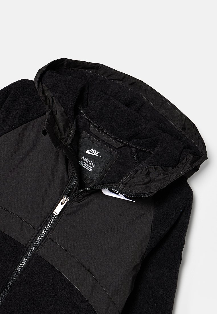 Giacca nera con cappuccio. Presenta una texture in pile, sezioni di tessuto liscio e una cerniera frontale. Include un logo Nike sul petto.