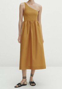 Robe jaune moutarde à une épaule avec un corsage smocké, une jupe fluide et un tissu texturé. Des sandales noires à brides complètent le look.