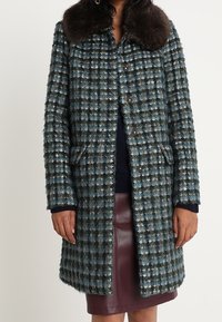 Ullmix-kappa i en teal- och brunrutigt mönster, med en faux fur-krage, knappstängning och sidofickor. Slät textur överlag.