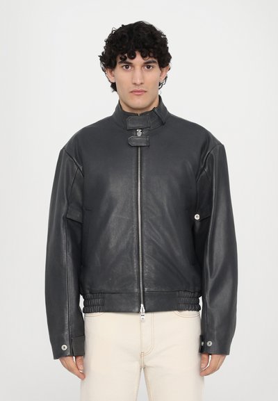 Veste en cuir noir avec un col haut, fermeture éclair et taille élastique. Coupe oversize avec des accents à boutons-pression sur les manches.
