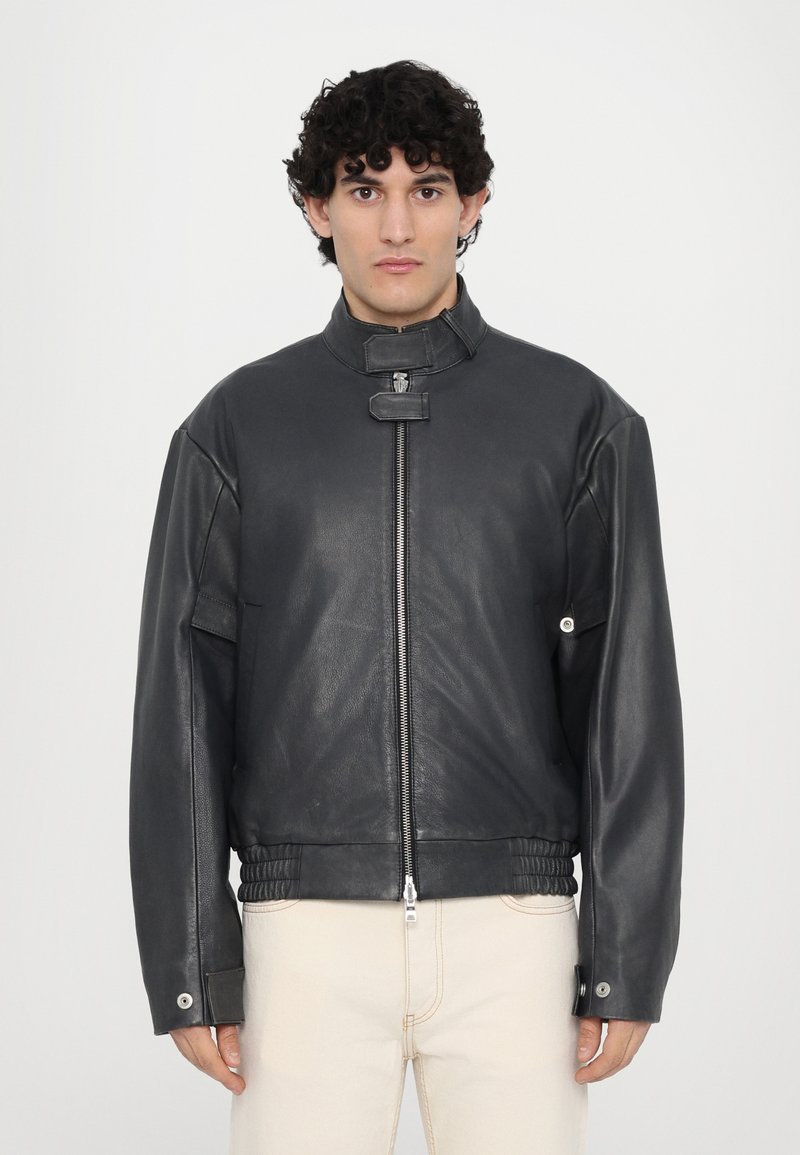 Veste en cuir noir avec un col haut, fermeture éclair et taille élastique. Coupe oversize avec des accents à boutons-pression sur les manches.