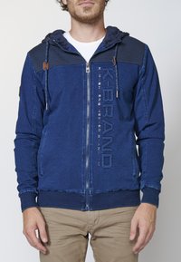 Navy blue zip-up hoodie in soft fabric with ribbed accents on shoulders, logo detail on front, and drawstring hood.