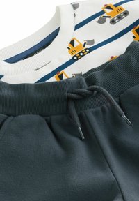 Twee-delige outfit: witte katoenen shirt met patronen van blauwe en gele bouwvoertuigen, gecombineerd met donkergrijze joggingbroek met een trekkoord.