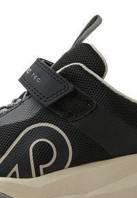 Scarpa sportiva nera con tomaia in rete, strap di velcro, logo prominente in crema, accenti testurizzati e suola imbottita beige.