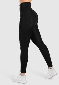 Leggings noirs taille haute en tissu extensible, dotés de poches latérales et d'un design sans coutures. Associés à des chaussures de sport blanches.