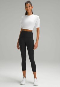 Top corto bianco a maniche corte abbinato a leggings neri a vita alta. Il modello completa il look con scarpe da ginnastica bianche.